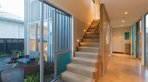 Interior view of a contemporary residential hallway with a carpeted staircase leading to an upper level. Full-height glazed doors and windows open toward a small courtyard or patio, while warm recessed lighting, timber-toned wall panels, and polished light flooring create a bright, refined circulation space.