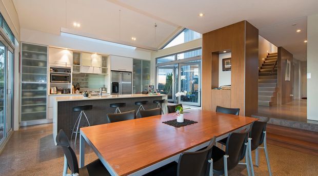 Open-plan contemporary kitchen and dining area with a large timber dining table, island bench with seating, stainless-steel appliances, extensive built-in cabinetry, and a timber-clad stair volume. The room has polished concrete flooring, high ceilings, clerestory glazing, and abundant natural light.