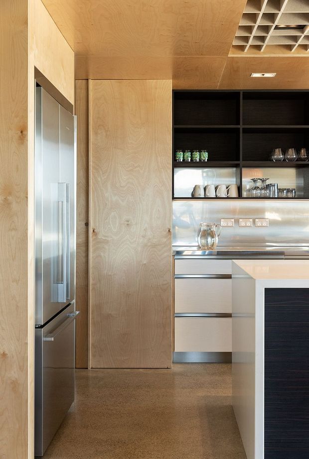 Compact contemporary kitchen with extensive light plywood wall and ceiling lining, integrated stainless-steel refrigeration and appliances, dark open shelving, metallic splashback, and a polished concrete or terrazzo-like floor. A white island or peninsula with dark textured paneling is visible in the foreground.