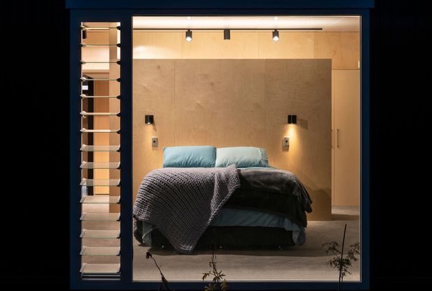 A compact, warmly lit bedroom viewed through a large dark-framed opening. The room features a low bed with layered blue-grey bedding, a textured throw, integrated bedside wall lights, pale timber or wood-look wall panelling, recessed ceiling spotlights, and built-in cabinetry along the right side. A narrow louvred or slatted screen is visible beside the opening. The overall design is restrained, contemporary, and intimate.