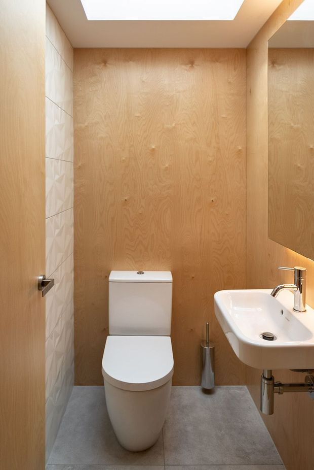 Compact contemporary bathroom with a wall-mounted white basin, toilet, large mirror, grey floor tiles, warm timber wall panelling, a textured white feature tile wall, and a rectangular ceiling skylight or lightwell.
