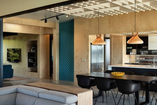 A contemporary open-plan kitchen and dining area viewed from the living room. The space features warm timber wall and cabinetry surfaces, a dark kitchen island and dining table, black dining chairs, stainless-steel appliances, a patterned teal feature wall, sculptural timber pendant lights, and a striking geometric coffered ceiling. A blue-toned living room sofa and armchair are visible in the foreground and adjacent area.