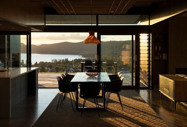 A sophisticated open-plan dining and kitchen interior with a dark, moody fit-out and expansive floor-to-ceiling glazing overlooking a lake and wooded hills. A rectangular stone-topped dining table with black chairs sits beneath a sculptural copper pendant, while dark kitchen cabinetry, timber ceiling battens, and a sideboard reinforce the refined contemporary atmosphere. Strong afternoon light casts linear shadows across the polished floor and rug.