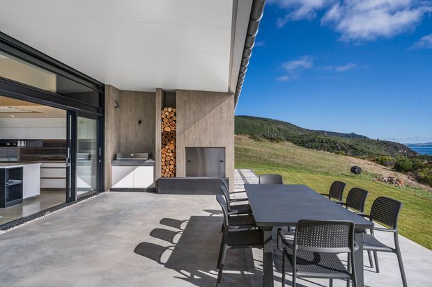 Modern outdoor terrace adjoining a glazed interior, with a concrete floor, dark-framed sliding doors, timber-toned wall cladding, built-in outdoor kitchen and fireplace niches, and a large outdoor dining setting. The terrace overlooks a grassy coastal hillside and ocean under a clear blue sky.
