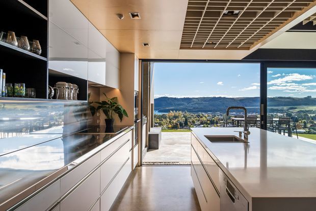 A contemporary kitchen with a long central island, integrated sink and tap, extensive white cabinetry, open black shelving, and a polished light benchtop. Warm timber ceiling panels and a decorative timber grid ceiling add contrast. Full-height glazing and an open glass door provide expansive views toward a hilly, semi-rural landscape and bring in abundant natural light.