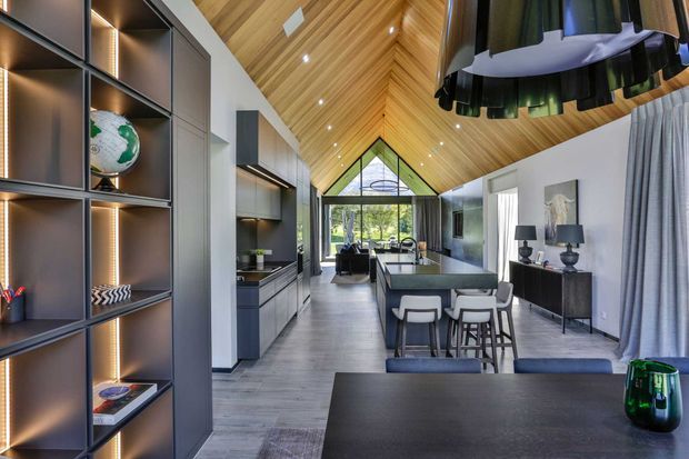 A spacious contemporary open-plan interior combining kitchen, dining and living areas. The room features a dramatic timber-lined pitched ceiling with recessed lighting, dark grey built-in cabinetry and shelving, a long island or breakfast bar, large glazed gable-end windows, a dining table in the foreground, and a sculptural dark pendant light. Neutral furnishings and grey flooring create a refined, modern atmosphere.
