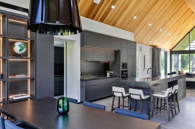 A spacious contemporary open-plan kitchen and dining area with dark grey full-height cabinetry, a large island with seating, integrated appliances, and a dark dining table. Warm timber slatted ceiling, expansive black-framed glazing, recessed lighting, and a sculptural black pendant create a refined contrast between warm wood and charcoal finishes.