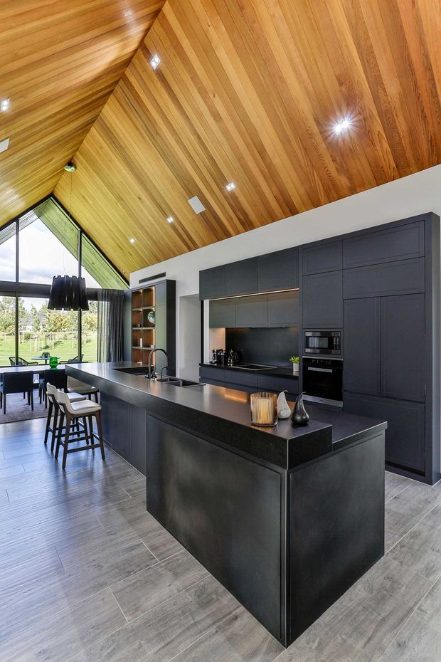 A contemporary open-plan kitchen and dining area with a large dark island, integrated matte charcoal cabinetry, black appliances, and a dark benchtop. The kitchen is framed by extensive glazing overlooking greenery and features a dramatic pitched ceiling lined in warm timber boards with recessed spotlights. Grey wood-look flooring, pendant lighting, stools, and a dining table complete the space.
