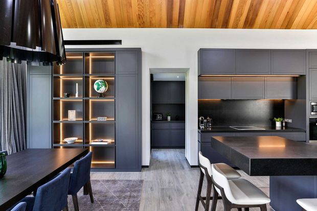 Contemporary open-plan kitchen and dining area with dark grey built-in cabinetry, a large dark stone island, integrated shelving with warm LED lighting, and a timber-lined ceiling. A compact dining table with upholstered chairs sits beside the kitchen, while a pantry or secondary storage area is visible through the central opening.