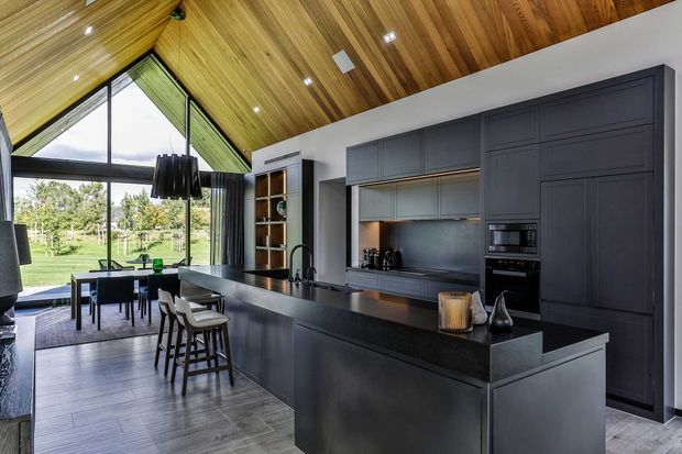 A dramatic open-plan kitchen and dining area with a large dark island, integrated dark cabinetry, black benchtops, and a stainless-steel-toned splashback. The room features a warm timber-lined pitched ceiling, recessed lighting, expansive triangular and full-height glazing, and a dining table overlooking a green outdoor setting.