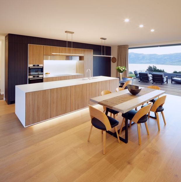 A bright open-plan kitchen and dining area with a large white waterfall island, integrated timber cabinetry, dark feature cabinetry, and a built-in oven wall. A timber dining table with sculptural wooden chairs sits beside the kitchen, opening to a furnished outdoor deck through expansive glazing with a water and mountain view.