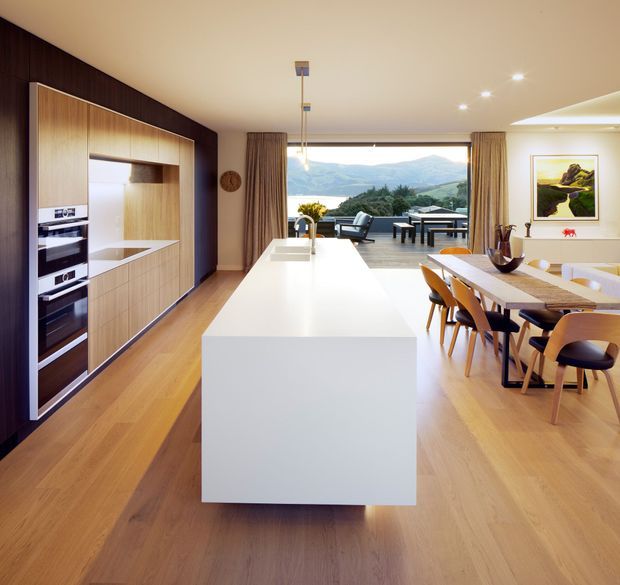 Open-plan contemporary kitchen and dining area with a large white island, integrated timber cabinetry and appliances, warm timber flooring, and a dining table beside full-height glazing. The glazing opens toward an outdoor terrace with a mountain landscape beyond. A dark timber feature wall contrasts with the light cabinetry and ceiling.