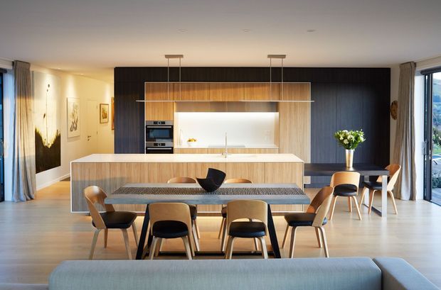 Open-plan contemporary interior featuring a kitchen, dining area and living space. The kitchen has a large timber island, integrated appliances, a dark timber feature wall, and a bright recessed splashback beneath pendant lighting. A timber dining table with upholstered chairs sits in the foreground, with floor-to-ceiling glazing and curtains along the sides.