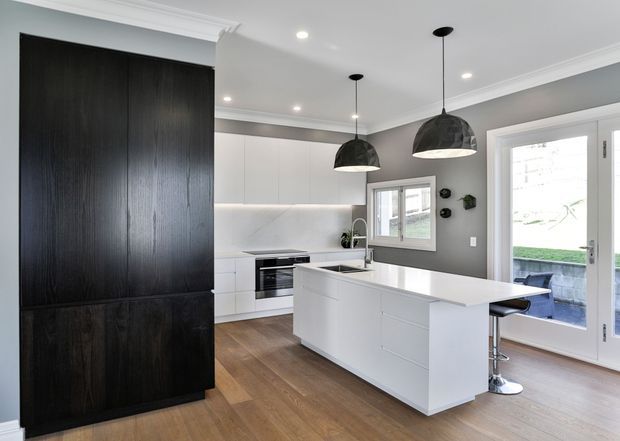 Bright contemporary kitchen with a large white island, integrated white cabinetry, dark timber feature storage wall, timber flooring, grey walls, pendant lights, and adjoining glazed doors opening to an outdoor area.