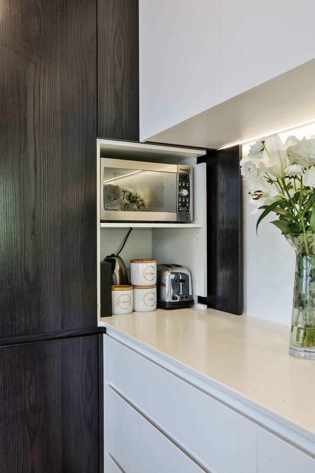 Close-up of a contemporary kitchen coffee station with integrated dark timber-grain cabinetry, white upper cabinets and drawers, a pale solid-surface benchtop, under-cabinet lighting, and a concealed appliance nook containing a microwave, kettle, toaster, and labelled canisters. A vase of flowers sits on the benchtop.