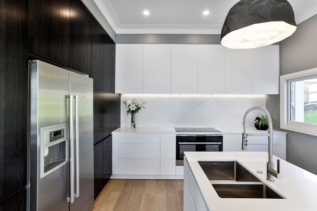 Bright contemporary kitchen featuring white handleless cabinetry, dark timber-look wall cabinetry, a white stone island with integrated double sink and gooseneck tap, stainless-steel appliances, timber flooring, and a sculptural black pendant light. A window provides natural light to the compact, polished space.