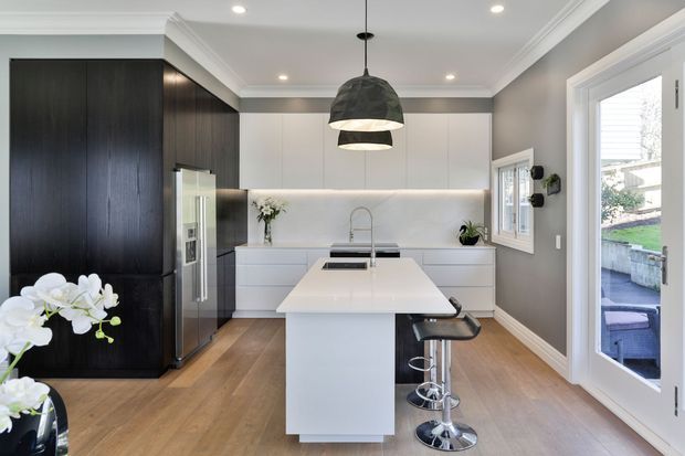 Contemporary kitchen with a large white island and integrated sink, dark full-height cabinetry and refrigerator surround, pale handleless wall and base cabinetry, timber flooring, grey walls, pendant lighting, and a glazed door opening to an outdoor area. The kitchen has a clean, refined, high-contrast material palette.