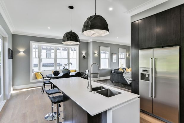 A contemporary open-plan kitchen and dining/living area with a large white island, integrated double sink and gooseneck tap, dark timber cabinetry, stainless-steel French-door refrigerator, and black pendant lights. Grey walls, pale timber flooring, large white-trimmed windows, and a grey sofa create a refined, monochromatic interior.