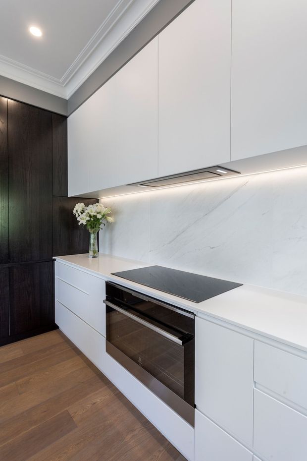 A refined contemporary kitchen featuring full-height white cabinetry, dark timber cabinetry, a white veined stone backsplash and benchtop, an integrated induction cooktop, built-in oven, under-cabinet lighting, and timber flooring. Decorative cornices add a subtle traditional architectural detail.