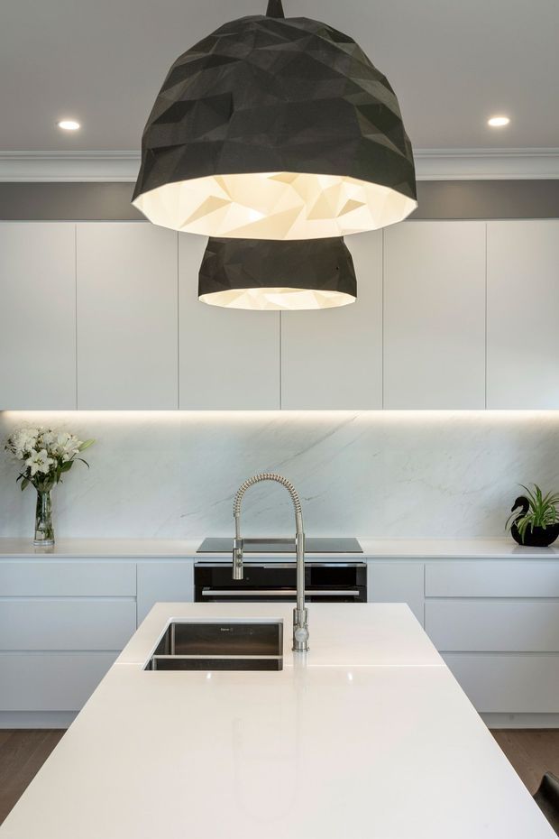 A contemporary kitchen centered on a large white island with an inset stainless-steel sink and tall gooseneck tap. The space features pale grey-white handleless cabinetry, a veined white stone splashback, integrated appliances, under-cabinet lighting, and two striking faceted black pendant lights with illuminated white interiors.