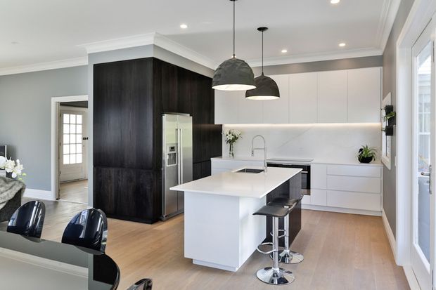 Bright contemporary kitchen with a large white island, integrated sink and seating, dark timber-look full-height cabinetry, white handleless cabinetry, pale timber flooring, and two dark pendant lights. A dining area is partially visible in the foreground, with a hallway and glazed exterior door beyond.