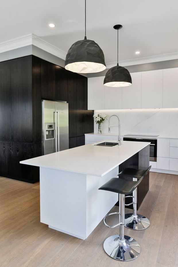 A contemporary kitchen featuring a large white island with integrated sink and waterfall-style sides, black bar stools, white cabinetry, a veined white splashback, and dark timber cabinetry surrounding a stainless-steel refrigerator. Two faceted black pendant lights hang above the island, with recessed ceiling lighting and light timber flooring completing the high-contrast scheme.