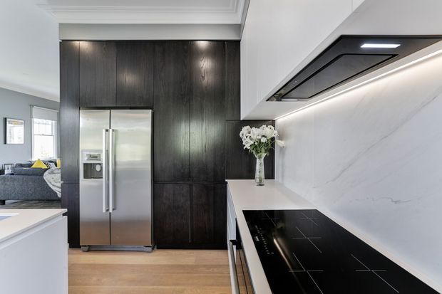 A bright contemporary kitchen with a long white stone benchtop, black induction cooktop, integrated rangehood, marble-look splashback, dark timber cabinetry, and a stainless-steel French-door refrigerator. A living area is visible beyond the kitchen, with recessed lighting and light timber flooring creating a refined, modern interior.