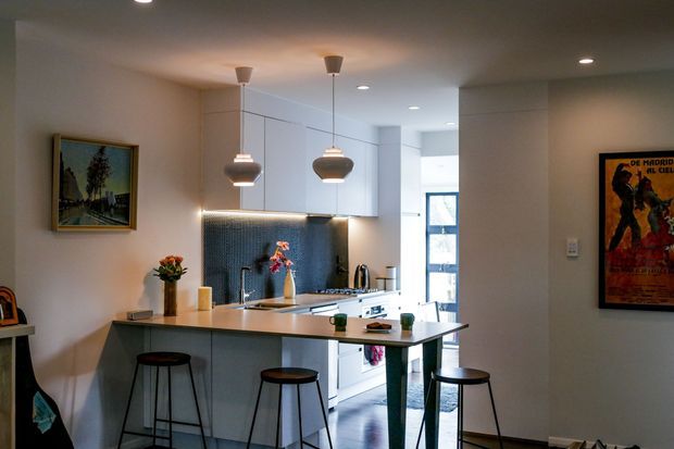 A compact, open-plan contemporary kitchen viewed from the adjoining living area. The space features white flat-panel cabinetry, a pale stone or engineered-stone island benchtop with integrated seating, a dark textured tiled splashback, stainless-steel appliances, pendant lights, and recessed ceiling lighting. Framed artwork and stools add warmth to the otherwise neutral interior.