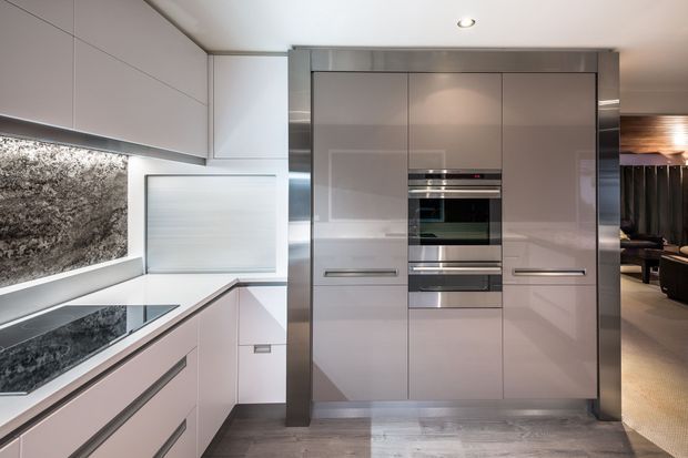 A sleek contemporary kitchen with glossy pale cabinetry, integrated stainless-steel ovens, a dark natural-stone splashback and benchtop, and streamlined handleless joinery. The compact kitchen opens toward a darker adjoining living or dining area.