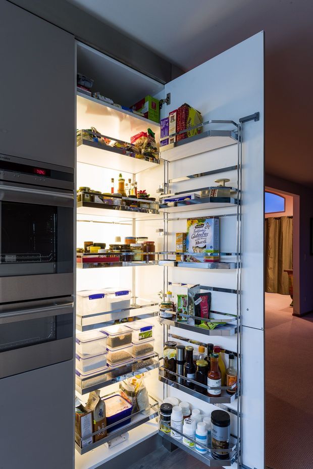 Open, fully stocked walk-in pantry with illuminated adjustable shelving and door-mounted racks, displaying dry goods, jars, bottles, containers, and packaged food. Integrated stainless-steel ovens are visible beside the pantry, with a glimpse of an adjacent room beyond.