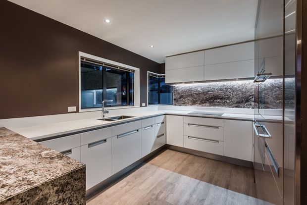 Contemporary L-shaped kitchen with sleek white handleless cabinetry, a dark brown painted feature wall, white benchtops, and a dramatic natural-stone-look splashback and peninsula. Integrated appliances, under-cabinet lighting, stainless fixtures, timber-look flooring, and a window over the sink complete the refined modern fit-out.