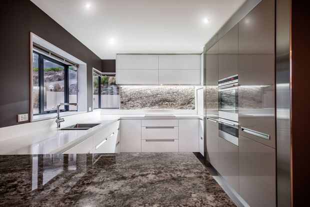 A contemporary L-shaped kitchen with white flat-panel cabinetry, integrated appliances, a large polished stone island and matching stone splashback. Dark brown walls and tall reflective cabinetry create a strong contrast, while a window and glazed door provide natural light and views outside.