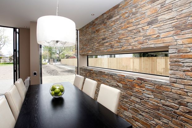 Contemporary dining room with a long black dining table, upholstered cream dining chairs, and a large white pendant light. A full-height rough stacked-stone feature wall dominates the room and incorporates a narrow horizontal glazed opening looking toward a timber fence. Large glazed doors on the left provide access and views outdoors.