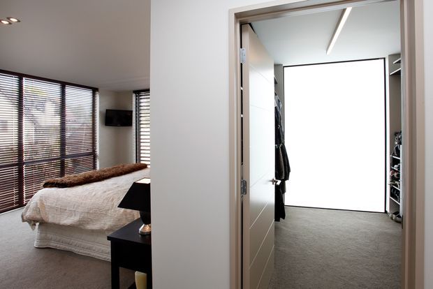 Interior view showing a contemporary bedroom on the left with a low upholstered bed, dark bedside table, and expansive timber venetian blinds, alongside an adjoining walk-in wardrobe with carpeted flooring, open shelving, hanging clothes, and a large bright opening or window.