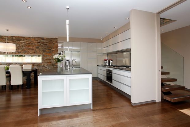 Open-plan contemporary kitchen and dining area with a white island, sleek white and grey built-in cabinetry, stainless-steel appliances, dark timber flooring, and a rustic exposed brick feature wall. A floating timber stairway is visible at the right.