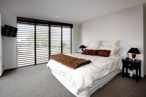 A bright, modern bedroom with a large upholstered bed dressed in white patterned bedding and brown textured throws and cushions. Full-height black-framed glazing with horizontal louvres spans one wall, while dark bedside tables and lamps provide contrast against the white walls and grey carpet.