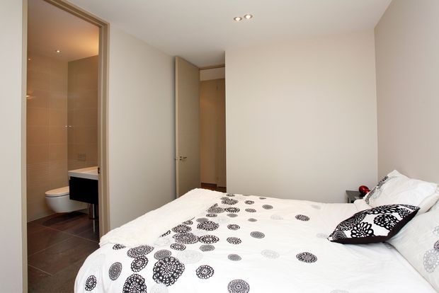 A bright, contemporary bedroom with a double bed dressed in white bedding and black patterned accents. An open doorway reveals an adjoining tiled bathroom with a toilet and dark vanity, while another door opens to a hallway or wardrobe area. Recessed ceiling lights and neutral painted walls create a clean, understated interior.