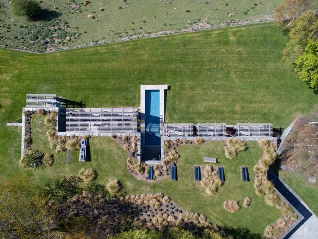 Aerial view of a landscaped residential outdoor area featuring a long rectangular swimming pool, extensive timber-toned slatted decking or screening, ornamental grasses, planted garden beds, and expansive lawns. A paved walkway borders the garden, with a natural rocky slope visible beyond.