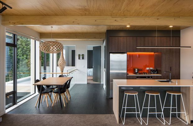 Open-plan contemporary kitchen and dining area with dark wood cabinetry, a vivid orange glass splashback, a light benchtop island with bar seating, and a timber-lined ceiling with exposed beam. Floor-to-ceiling glazing brings in abundant natural light beside a compact dining table and sculptural woven pendant lights.