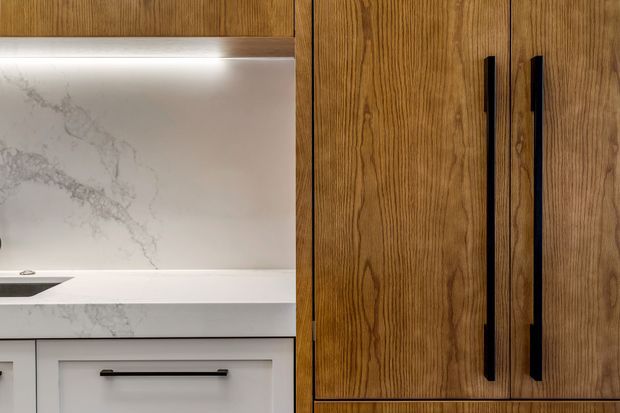 Close-up view of refined kitchen cabinetry, featuring warm vertical-grain timber veneer doors with long matte-black pulls, pale cabinetry below, and a white marble-look stone splashback illuminated by concealed under-cabinet lighting. The image focuses on fixed joinery and material detailing.