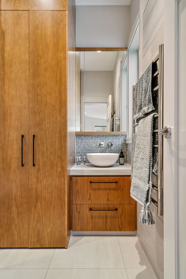 A compact contemporary bathroom viewed from the doorway, featuring full-height warm timber cabinetry and vanity drawers, a white vessel basin on a pale stone benchtop, small grey mosaic tile splashback, large wall mirror, and a wall-mounted chrome towel warmer with textured towels. Pale large-format floor tiles and light neutral walls create a clean, refined finish.