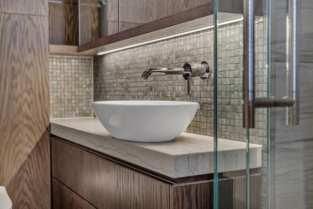 Close-up of a refined bathroom vanity with a white vessel basin set on a pale stone benchtop, dark timber-grain cabinetry, and a wall-mounted chrome tap. Small textured square tiles form the splashback, with integrated under-shelf lighting and a frameless glass shower enclosure visible at right.