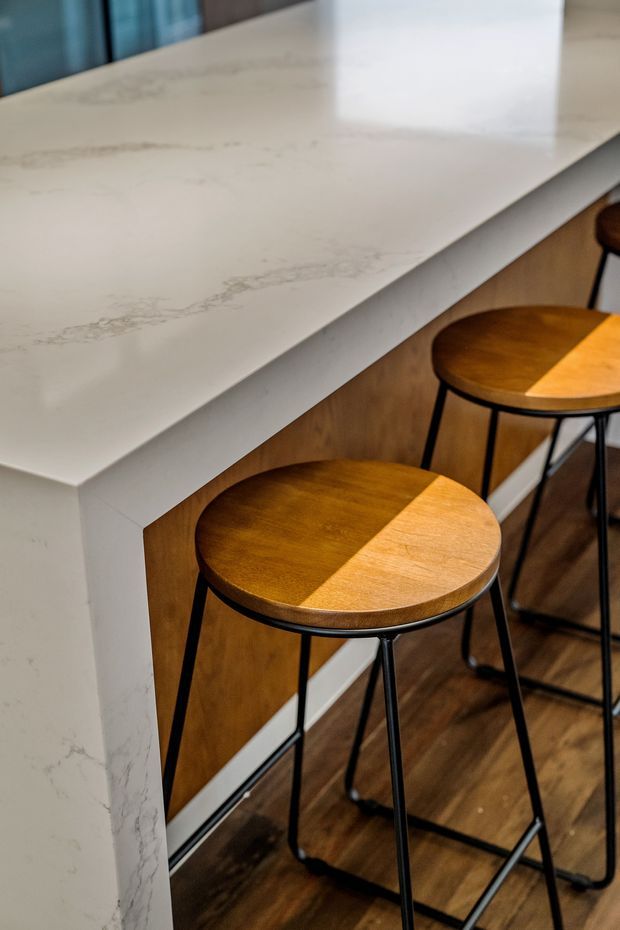 Close-up view of a kitchen or entertaining counter with a white veined stone benchtop, warm timber panelled base, and round timber stools with slim black metal legs. Dark timber flooring and a bright, contemporary fit-out are visible.