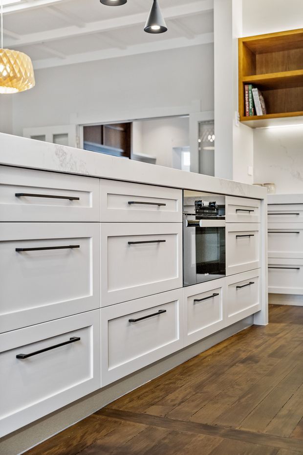 Bright, refined kitchen featuring extensive white Shaker-style cabinetry with dark linear handles, a pale marble-look benchtop, integrated oven, timber herringbone flooring, open timber shelving, and pendant lighting. A large interior opening and reflective surfaces create a sense of connection and spaciousness.