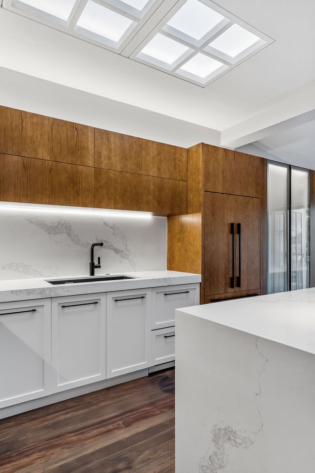 Bright contemporary kitchen featuring white lower cabinetry, warm wood-grain upper cabinets and tall pantry-style joinery, a white veined stone benchtop and waterfall island, integrated sink, dark timber flooring, and a large geometric skylight. Black tapware and handles provide contrast, while glazed doors bring additional daylight into the space.