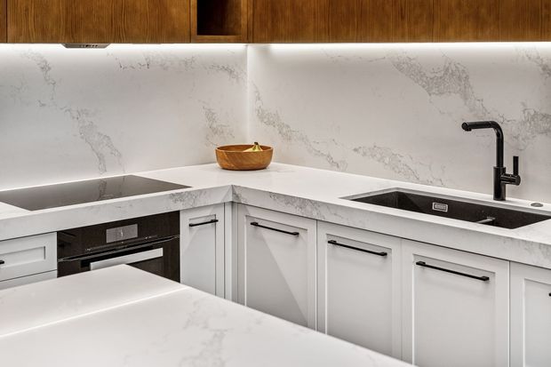 A contemporary L-shaped kitchen with white marble-look benchtops and splashback, white shaker-style cabinetry with black bar handles, integrated appliances, a black undermount sink and matte black tapware. Warm timber overhead cabinets and concealed LED strip lighting add contrast.