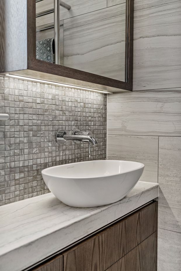Close-up of a refined bathroom vanity featuring a white vessel basin on a pale stone countertop, dark timber cabinetry, a wall-mounted chrome tap, large pale stone-look wall tiles, and a small textured square-tile splashback beneath a mirrored cabinet.