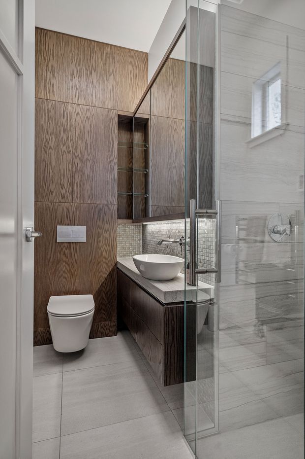 A compact contemporary bathroom featuring large grey floor and wall tiles, a wall-hung toilet, floating dark timber vanity with a white vessel basin, mirrored storage, mosaic tile splashback, and a frameless glass shower enclosure. Vertical timber wall panelling adds warmth and contrast.