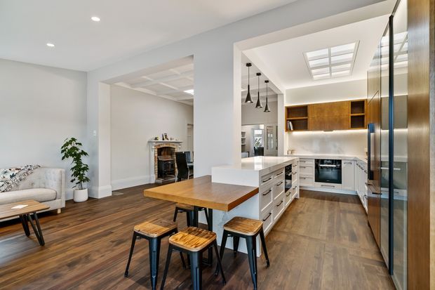 Bright open-plan kitchen and dining area with dark timber flooring, a white island with integrated cabinetry and seating, warm timber dining table, pendant lights, and wood-toned kitchen cabinetry. A living area and fireplace are visible through the wide opening.