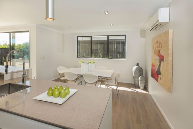 Bright open-plan kitchen and dining area with a large pale stone island, integrated sink and gooseneck tap, white cabinetry, timber-look flooring, and a contemporary dining setting. Neutral walls, recessed lighting, pendant lighting, artwork, and decorative vessels create a clean, airy interior.